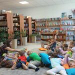 Grupa dzieci leży na poduszkach w kolorowej bibliotece, słuchając kobiety. W tle półki z książkami, rośliny i pluszowe misie. Na podłodze jasny dywan.