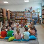 Grupa dzieci siedzi na podłodze w bibliotece na kolorowych poduszkach. W tle regały z książkami i rośliny. Prowadząca kobieta uczestniczy w zajęciach.