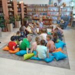 Dzieci siedzą na kolorowych poduszkach w jasnej bibliotece. Tło to półki z książkami i pluszowe misie. Rośliny dodają zieleni.