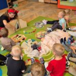 Dzieci siedzą w kręgu na kolorowym dywanie w przedszkolu. Kobieta z chustką na głowie pokazuje im pacynki. W tle są szafki i zabawki.