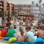 Grupa dzieci siedzi na kolorowych poduszkach w bibliotece. Przed nimi młoda kobieta czyta książkę. W tle regały z kolorowymi książkami.