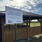 Drewniana altana na tle zielonego pola i błękitnego nieba. Na pierwszym planie biała tablica z czerwonym napisem. Altana ma ciemny dach.