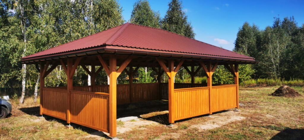 Drewniana altana z brązowym dachem stoi na trawie. Otoczona jest drzewami w tle. Jasne, słoneczne niebo dodaje kontrastu.
