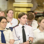 Na zdjęciu widać grupę młodych osób w białych uniformach siedzących w pomieszczeniu z beżowym tłem. Wszystkie osoby mają skupiony wyraz twarzy. W tle widać kolumnę i rozmyte ściany.