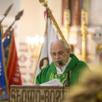 Na zdjęciu starszy mężczyzna w zielonym ornacie przemawia przy mównicy w kościele. W tle widać kolorowe sztandary. Pomieszczenie jest jasno oświetlone.
