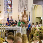 Kościół pełen wiernych, kapłan w zielonej szacie przy ambonie mówi kazanie. W tle kolorowe witraże i sztandary.