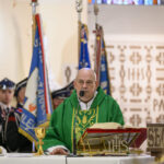 Kapłan w zielonym ornacie prowadzi mszę w kościele. Na pierwszym planie leży chleb i złoty kielich. W tle widoczni strażacy i sztandary.