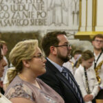 Grupa ludzi siedzi w kościele, w tle napis na ścianie. Dominują kolory białe i złote. Ludzie skupieni na wydarzeniu.