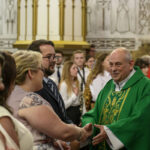 Nie wiem, kto jest na zdjęciu. Duchowny w zielonej szacie ściska dłonie wiernych. W tle malowidła i zgromadzenie ludzi.