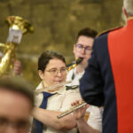 Osoby grające na instrumentach dętych w formalnych strojach. Dominują złote i czerwone kolory. Tło jest rozmyte i ciemne.