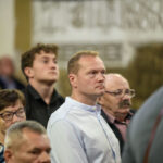 Na zdjęciu widać grupę osób stojących w pomieszczeniu. Dominują stonowane kolory ubrań i neutralne tło. Ludzie są skupieni, patrzą w jednym kierunku.