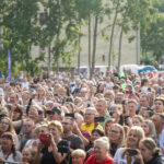 Zdjęcie przedstawia tłum ludzi na zewnątrz, w słoneczny dzień. W tle widoczne są drzewa i namiot zielonego koloru. Większość osób trzyma smartfony, wyglądają na rozradowane.