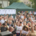 Grupa ludzi na plenerowym wydarzeniu, robiących zdjęcia telefonami. W tle zielone namioty i drzewa. Atmosfera radosna i słoneczna.