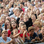 Grupa ludzi na koncercie, z uśmiechami i telefonami, ubrani w kolorowe stroje. Tło jest rozmyte. Widać żywą atmosferę.