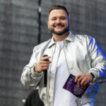 Osoba w srebrnej kurtce trzyma mikrofon na tle ciemnoszarej sceny. W dłoni ma fioletową kartkę. Uśmiecha się.