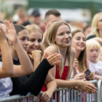 Grupa osób stoi za barierką na koncercie na świeżym powietrzu. Widać uśmiechnięte twarze, radosną atmosferę i różnorodne kolory ubrań. Tło jest rozmyte, sugerując środowisko festiwalowe.