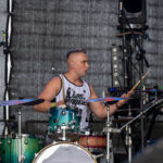 Perkusista w białej koszulce gra na zielono-brązowej perkusji. W tle ciemna scena z mikrofonem i sprzętem. atmosfera koncertu.