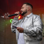 Mężczyzna w srebrnej kurtce trzyma mikrofon. Tło jest ciemne z kolorowymi światłami. Scena koncertowa, dynamiczna atmosfera.