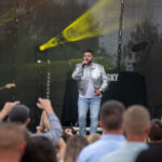 Scena koncertowa z wokalistą w srebrnej kurtce, gitarzystą obok i tłumem fanów na pierwszym planie. Tło czarne, oświetlone żółtym światłem.