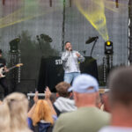 Na scenie mężczyzna śpiewa, ubrany w srebrną kurtkę. Obok gitarzysta, w tle ciemna scenografia i kolorowe światła. Publiczność rozentuzjazmowana, widać uniesione ręce.