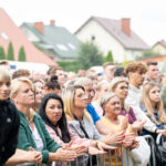 Grupa ludzi na wydarzeniu na świeżym powietrzu, stojących za barierkami. Kolorowe domy w tle. Uczestnicy są uważni i skupieni.