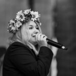 Osoba w wieńcu z kwiatów śpiewa do mikrofonu. Tło rozmyte, czarno-białe zdjęcie. Trzyma kartkę z tekstem.