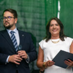 Dwie osoby na scenie, mężczyzna w garniturze z mikrofonem, kobieta w białej sukience z teczką. Tło zielone.