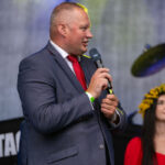 Mężczyzna w garniturze i czerwonym krawacie mówi do mikrofonu. W tle rozmyte światła sceniczne i kobieta w czerwieni z wiankiem. Dominują kolory niebieski, czerwony i żółty.