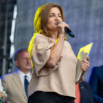 Na zdjęciu kobieta w beżowej bluzce przemawia do mikrofonu, trzymając dokumenty. W tle grupa osób formalnie ubranych. Scena ma szare tło.