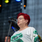 Osoba z czerwonymi włosami mówi do mikrofonu na scenie. Ma białą bluzkę z zielonym wzorem. Tło jest szare z kolorowymi światłami.