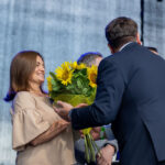 Na zdjęciu kobieta z uśmiechem odbiera bukiet słoneczników od mężczyzny. W tle ciemnoszara kurtyna. Obok stoją osoby w niebieskich koszulach.