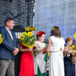 Grupa ludzi na scenie, wręczających sobie kwiaty i prezenty. W tle szare zasłony, dominują kolory zielony i żółty. Uroczysta atmosfera.