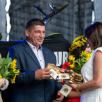 Na zdjęciu grupa ludzi przy scenie dekoruje kogoś kwiatami. Dominują kolory żółty z słoneczników oraz niebieski. Tło jest ciemne.