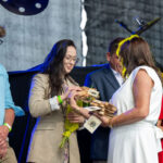 Na zdjęciu grupa osób na scenie. Kobieta w jasnym ubraniu przekazuje drewniane tabliczki. W tle widać osoby z bukietami słoneczników.