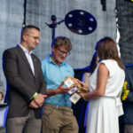 Na zdjęciu grupa ludzi na scenie, wręcza nagrody. Mężczyzna w niebieskiej koszuli trzyma drewniane trofea. Tło ciemne, z elementami sprzętu muzycznego.