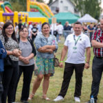 Sześć osób stoi na trawie przed kolorowymi dmuchańcami. Tło to jasne namioty i ogrodzenie. Na zdjęciu dominują kolory: żółty i zielony.