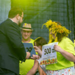 Na scenie mężczyzna przekazuje kobiecie nagrodę pieniężną. Kobieta nosi wianek z żółtych kwiatów i jasnożółtą koszulkę. W tle ludzie w strojach ludowych na ciemnym tle.