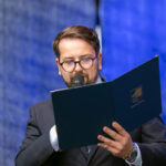 Mężczyzna w okularach przemawia do mikrofonu, trzymając niebieską teczkę. Tło w kolorze niebieskim. Ubrany w ciemny garnitur.