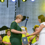 Na zdjęciu scena z osobami odbierającymi nagrody. Jedna osoba w zielonym, druga w białym, tło szare z oświetleniem. Grupka ludzi w jasnych koszulkach.