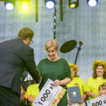 Na scenie kobieta odbiera nagrodę w wysokości 1000 zł w otoczeniu ludzi w jaskrawych koszulkach. Tło jest szare, a scena oświetlona żółtymi światłami. Obok stoi kobieta z mikrofonem.