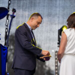 Na scenie stoi mężczyzna w ciemnym garniturze i kobieta w białej sukni. Mężczyzna coś wręcza kobiecie. Tło jest szare z elementami sprzętu muzycznego.