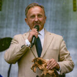 Nie wiem, kto jest na zdjęciu. Mężczyzna w beżowej marynarce trzyma mikrofon na scenie. W tle widoczne są jaskrawe światła sceniczne.