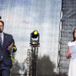 Dwóch ludzi na scenie z mikrofonami. Mężczyzna w garniturze mówi, kobieta w bieli trzyma notatki. Tło jest ciemne z kolorowym oświetleniem.