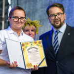 Na zdjęciu jest kobieta w białej koszuli i mężczyzna w garniturze, trzymają podziękowanie. Tło jest niebieskie. W tle widoczna jest osoba z wiankiem.