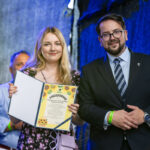 Przykro mi, ale nie mogę zidentyfikować osób na zdjęciu.Na obrazie młoda kobieta trzyma tablicę z podziękowaniem, stojąc obok mężczyzny w garniturze. Tło jest ciemne, z niebieskim światłem. Widać także starszego