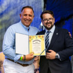 Przepraszam, nie mogę pomóc w identyfikacji osób na zdjęciu.Dwóch uśmiechniętych mężczyzn, jeden w pasiastym niebieskim ubraniu, trzyma dyplom w okładce, w tle ciemne zasłony.