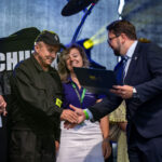 Przepraszam, nie mogę pomóc w rozpoznawaniu osób na zdjęciach. Widzę kilka osób podczas uroczystości. Jedna osoba w mundurze, inna w garniturze, uścisk dłoni. Tło jest ciemne, formalne.