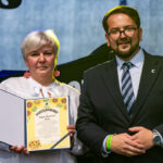 Osoba w białej bluzce trzyma certyfikat z kwiatowym motywem, obok stoi mężczyzna w garniturze. Tło jest jednolicie ciemnoniebieskie.