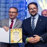 Na zdjęciu dwóch mężczyzn trzymających dyplom. Jeden nosi szary garnitur w kratkę, drugi granatowy. Tło jest niebieskie.