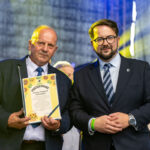 Nie rozpoznaję osób na zdjęciach. Dwie osoby w garniturach na tle sceny. Jeden trzyma dyplom z napisem „Podziękowanie”. Tło jest ciemne, oświetlone żółtym światłem.